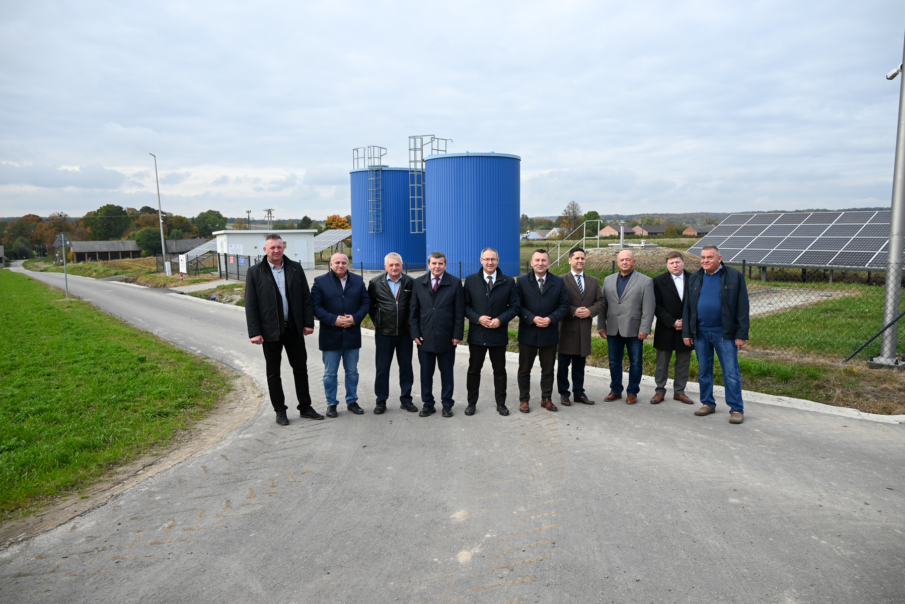Dziesięciu mężczyzn w ciemnych płaszczach i kurtkach stoi na szarej drodze, za nimi trzy niebieskie cylindryczne zbiorniki i panele słoneczne na tle zielonej trawy i zachmurzonego nieba.