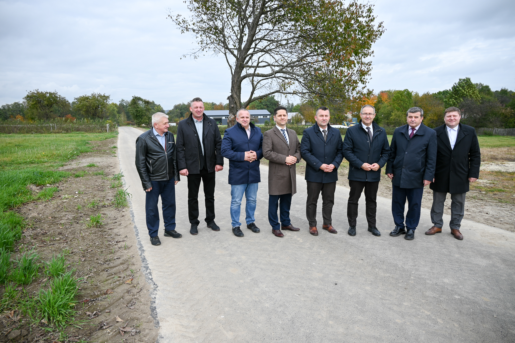 Osiem mężczyzn w jesiennych płaszczach stoi na szerokiej, szarej drodze, za nimi drzewo, zielona trawa i pola pod pochmurnym niebem.