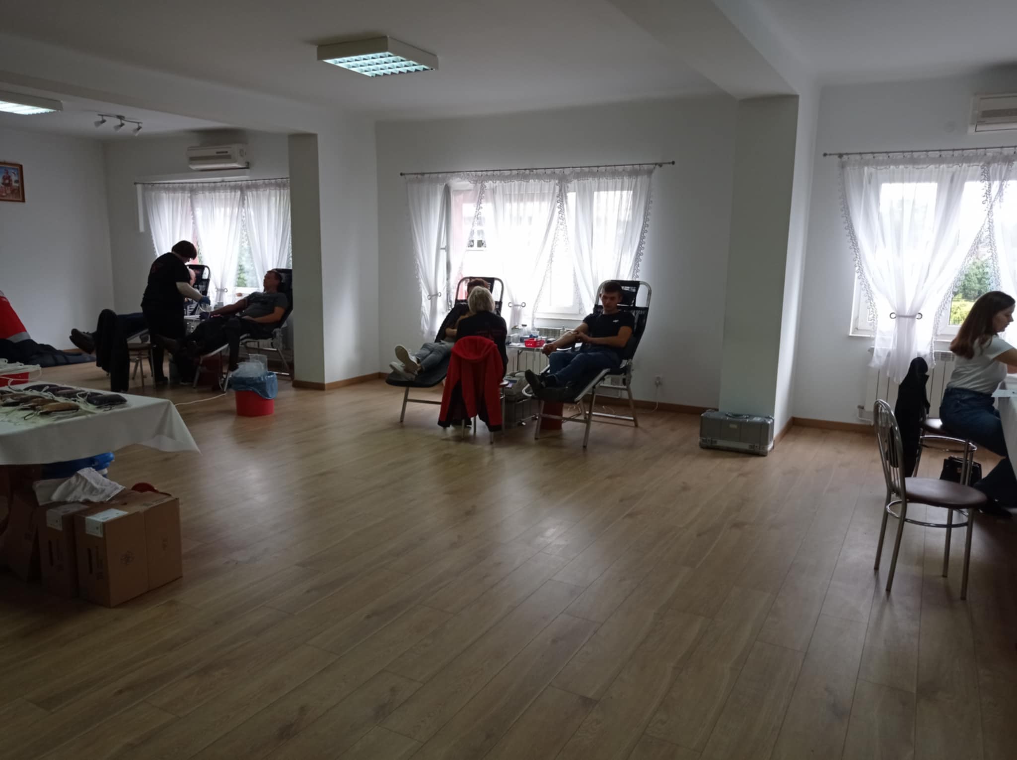 Wnętrze jasnego pomieszczenia z drewnianą podłogą, białe ściany, trzy okna z białymi firankami, kilka osób siedzących na składanych fotelach, stół z kartonami i sprzętem medycznym.