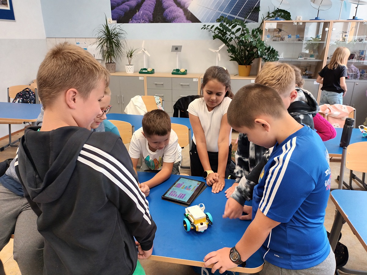 Grupa dzieci wokół niebieskiego stołu, na którym stoi mały robot i leży tablet z kolorowym ekranem, w tle szafki, rośliny i obraz ścienny z polem lawendy.