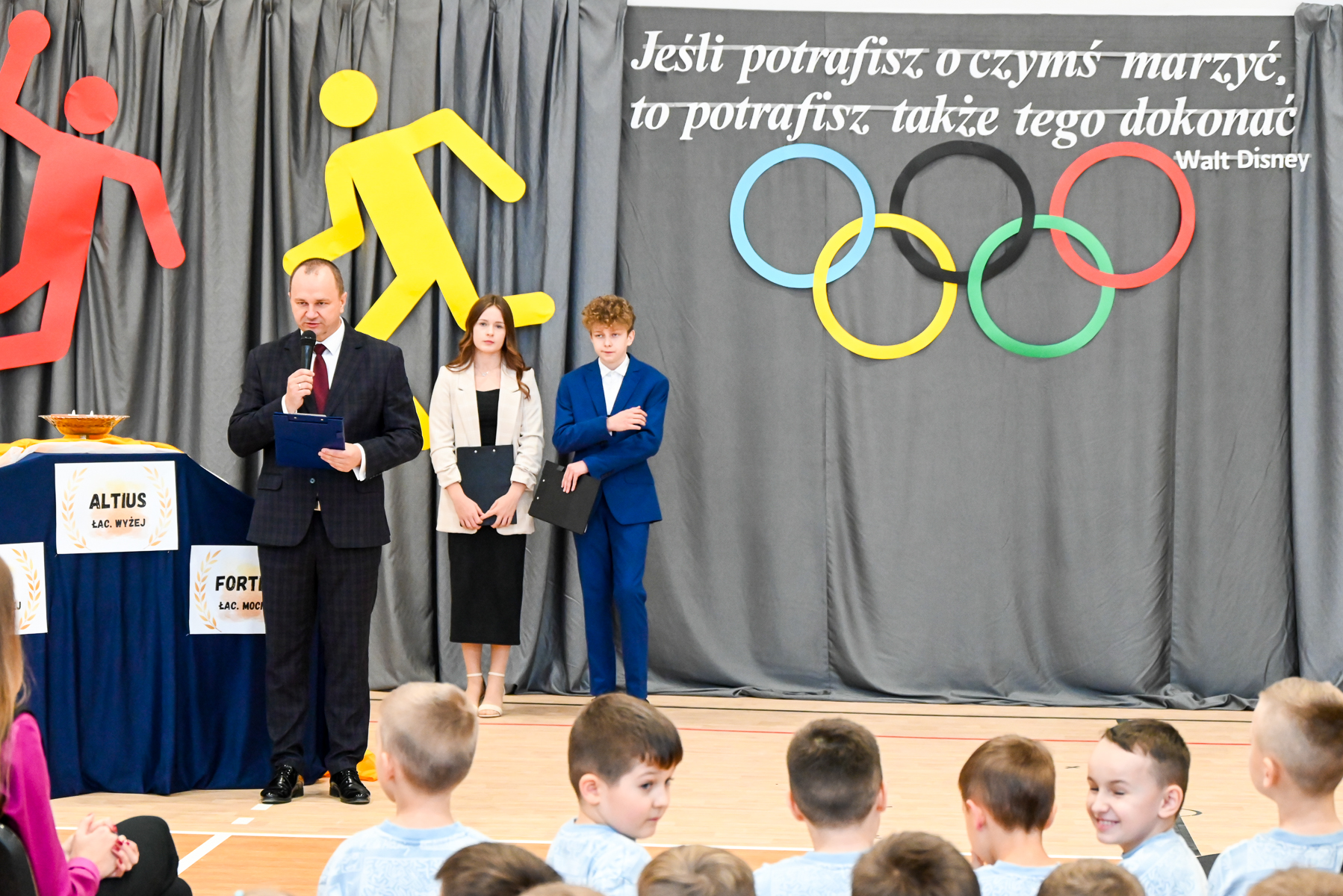 Mężczyzna przemawia do mikrofonu, obok stoją młoda kobieta i młody mężczyzna, za nimi szare zasłony z kolorowymi sylwetkami sportowców i napis Walt Disney z cytatem, dzieci siedzą na pierwszym planie.