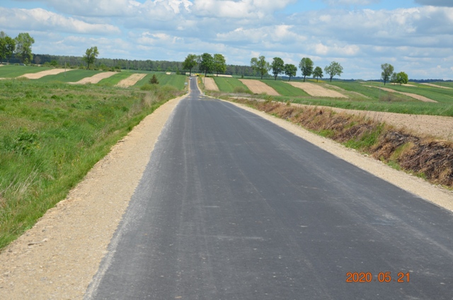 Asfaltowa droga na tle zielonych i żółtych pól, po bokach pobocze z ziemi, w oddali drzewa pod niebieskim niebem z chmurami.