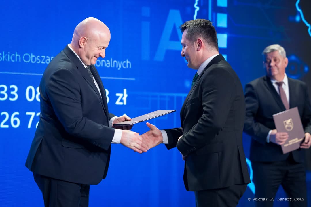 Dwaj mężczyźni w garniturach podają sobie ręce na tle niebieskiego ekranu, trzeci mężczyzna w garniturze trzyma teczkę i stoi z tyłu.