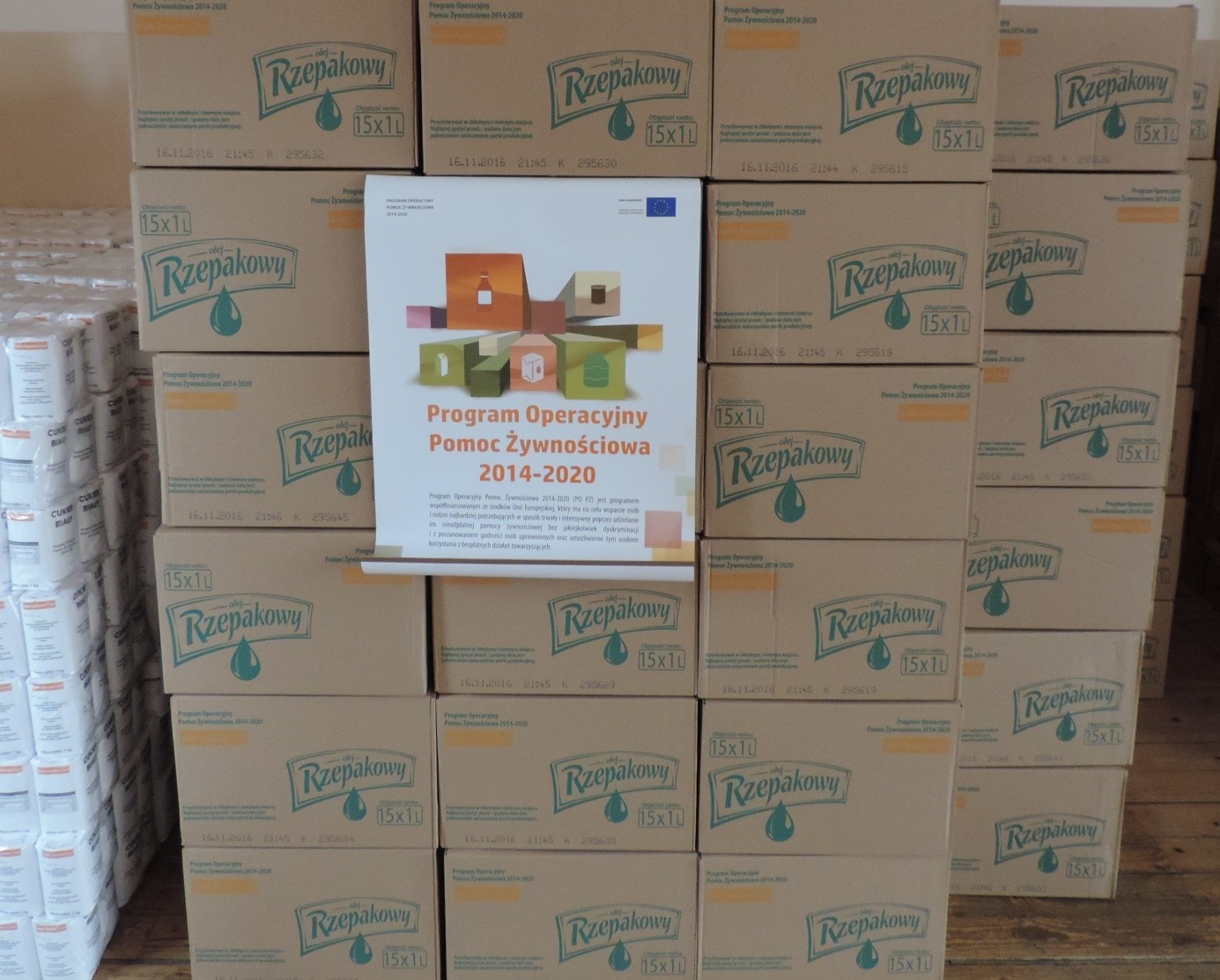 Stosy brązowych kartonów z napisem "olej Rzepakowy" oraz biały plakat z napisem "Program Operacyjny Pomoc Żywnościowa 2014-2020".