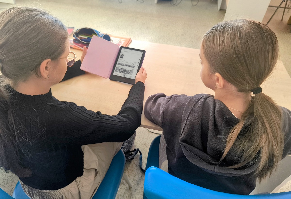 Dwie dziewczynki z jasnymi włosami związanymi w kucyki siedzą przy drewnianym stole na niebieskich krzesłach, jedna trzyma e-czytnik z różową okładką.