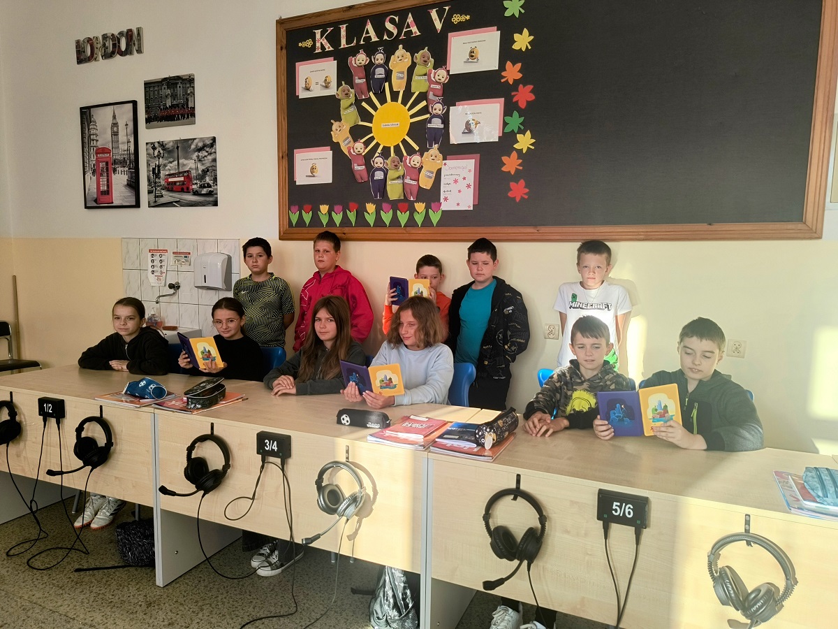 Grupa dzieci siedzących i stojących przy biurkach z słuchawkami, tablica z napisem "KLASA V" i kolorowymi dekoracjami, zdjęcia Londynu na ścianie.