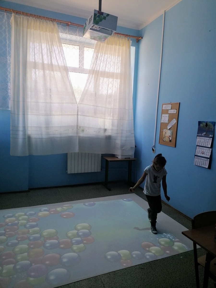 Pokój z jasnoniebieskimi ścianami, dużym oknem z białymi zasłonami, stołem w rogu i dzieckiem na podłodze z kolorowymi, okrągłymi kształtami wyświetlanymi przez sufitowy projektor.