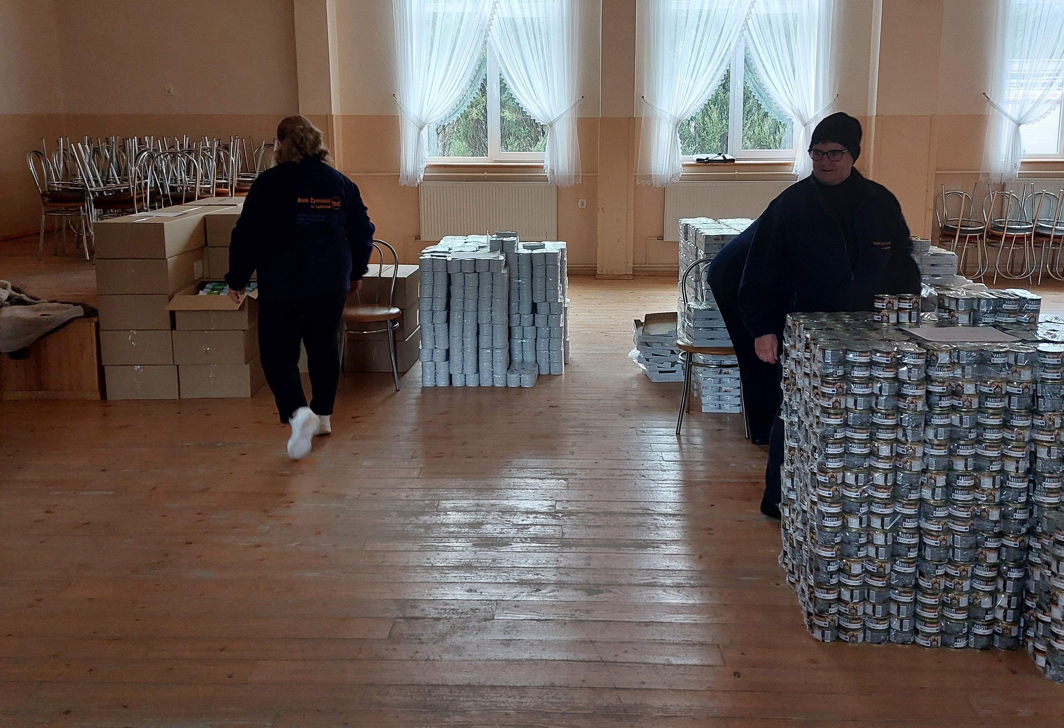 Drewniana podłoga, dwie kobiety w ciemnych ubraniach, sterty białych i ciemnych puszek oraz kartonów, jasne zasłony i okna z widokiem na zieleń.