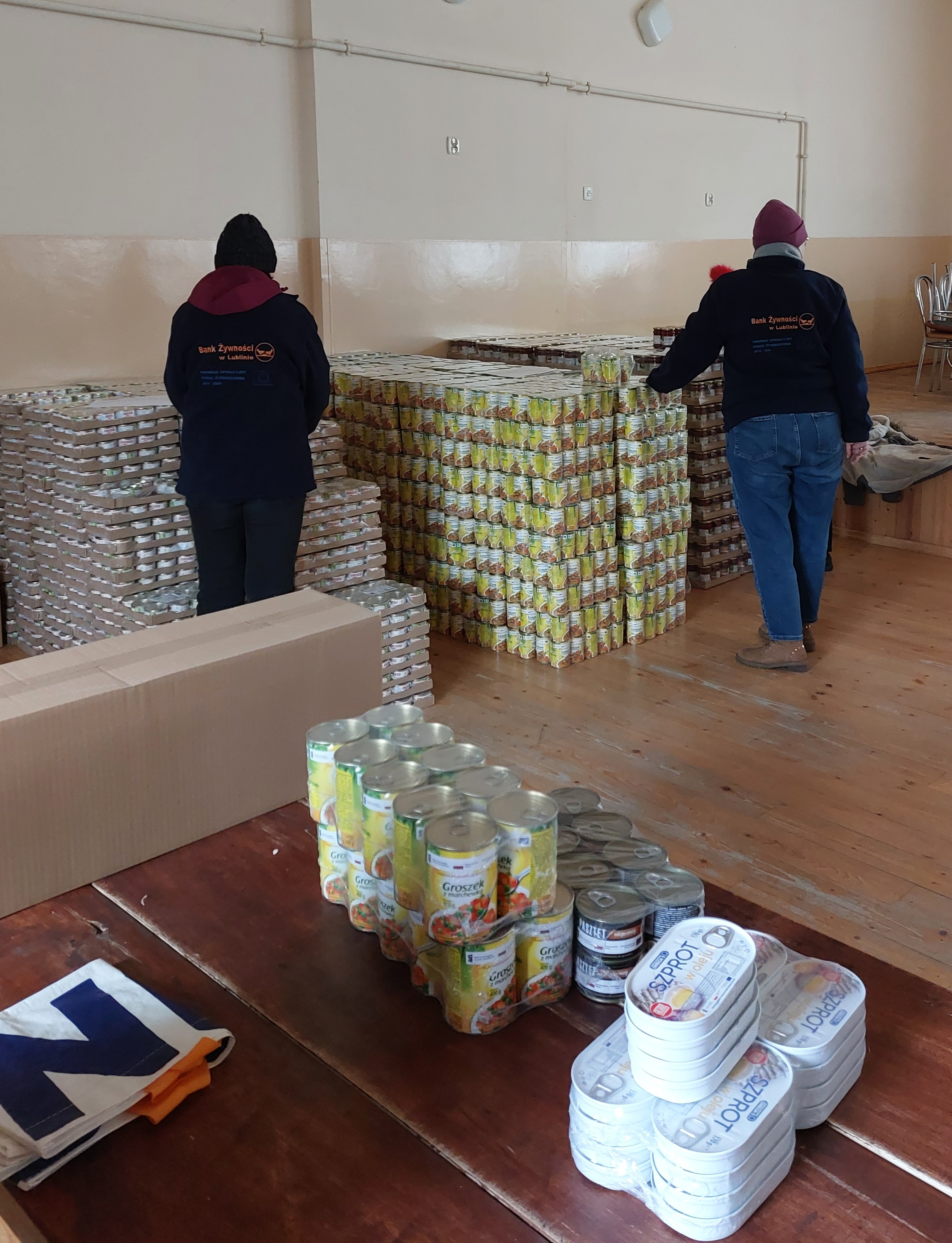 Dwie osoby w ciemnych kurtkach z napisem Bank Żywności w Lublinie stoją przy dużych stosach puszek na drewnianej podłodze w jasnym pomieszczeniu.