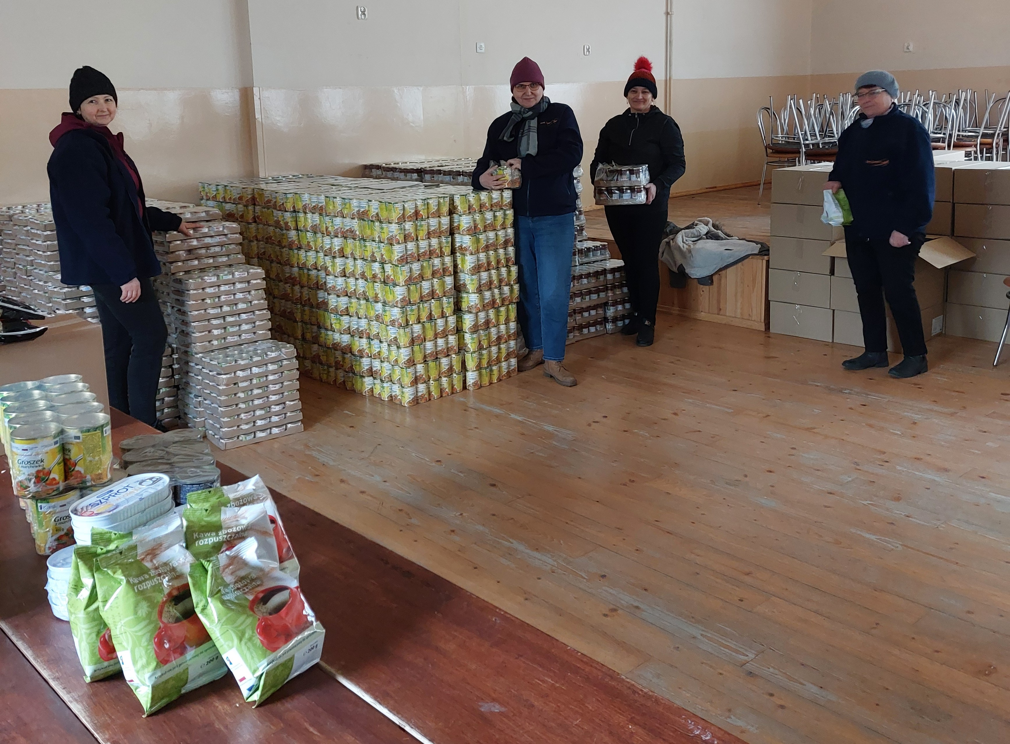 Cztery osoby w czapkach i kurtkach stoją w pomieszczeniu z drewnianą podłogą, wokół nich stosy puszek i kartonów z jedzeniem oraz paczki kawy na stole.