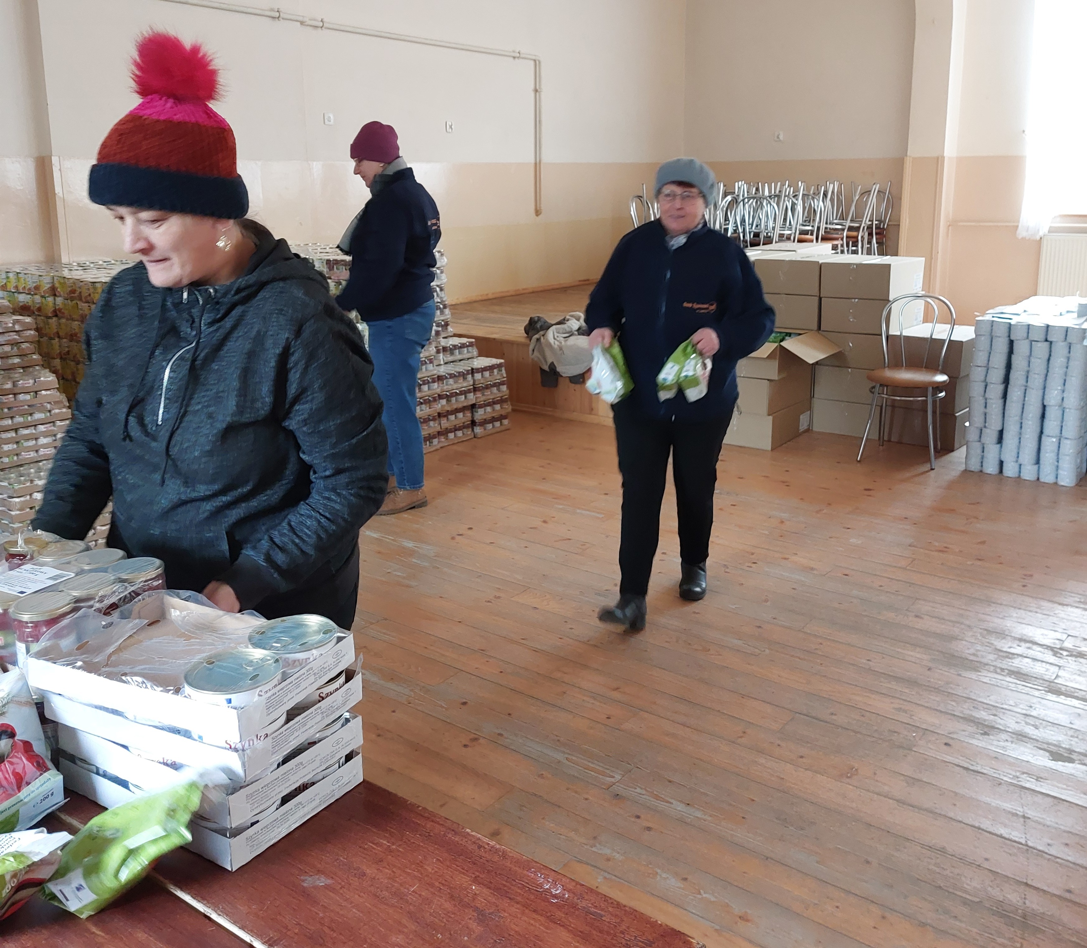 Trzy osoby w ciepłych ubraniach w pomieszczeniu z drewnianą podłogą, kartonami, stosami puszek i krzesłami na podwyższeniu.