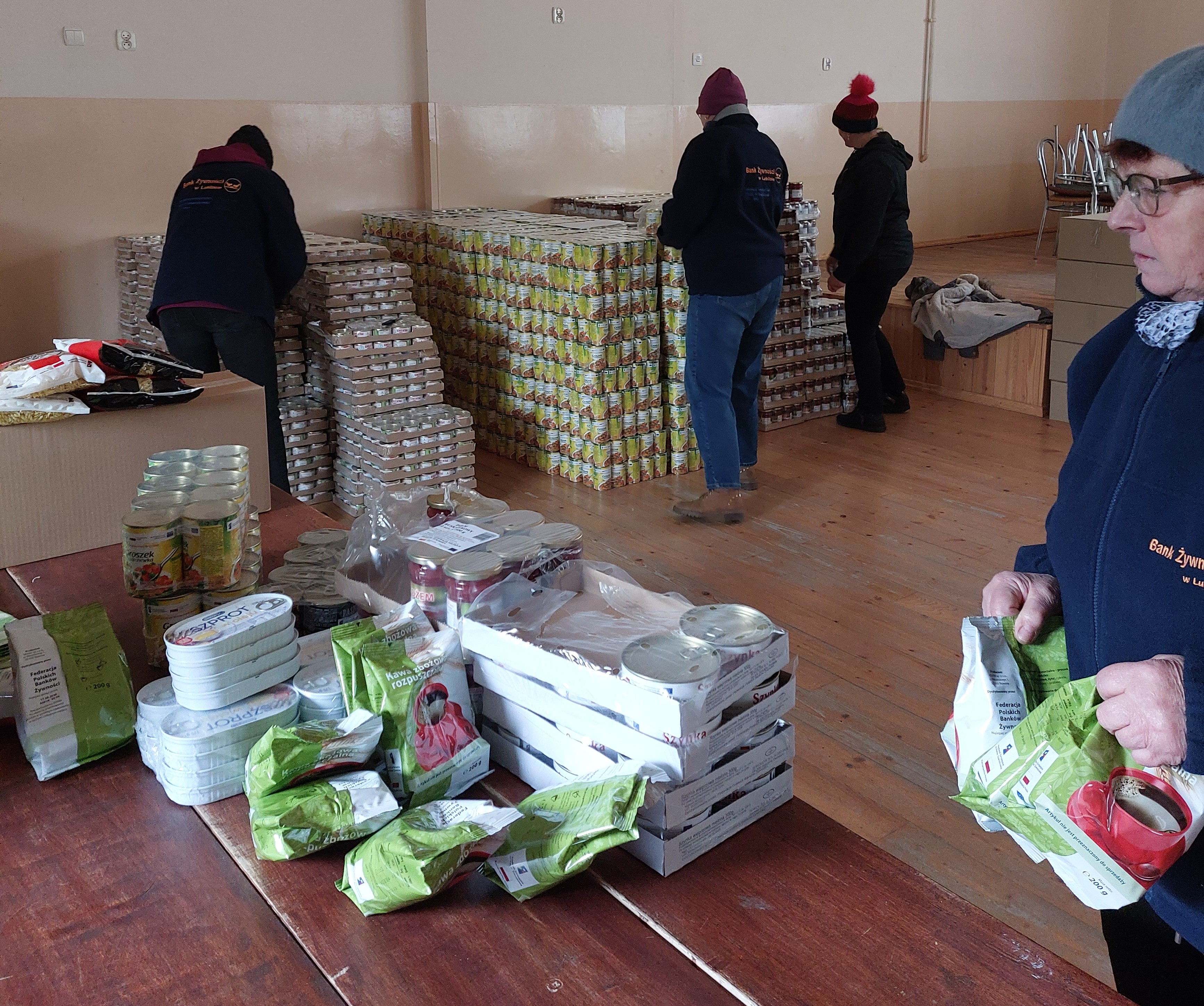 Cztery osoby w ciemnych ubraniach i czapkach obok stołu i dużych stosów puszek oraz paczek z żywnością w jasnym pomieszczeniu z drewnianą podłogą.