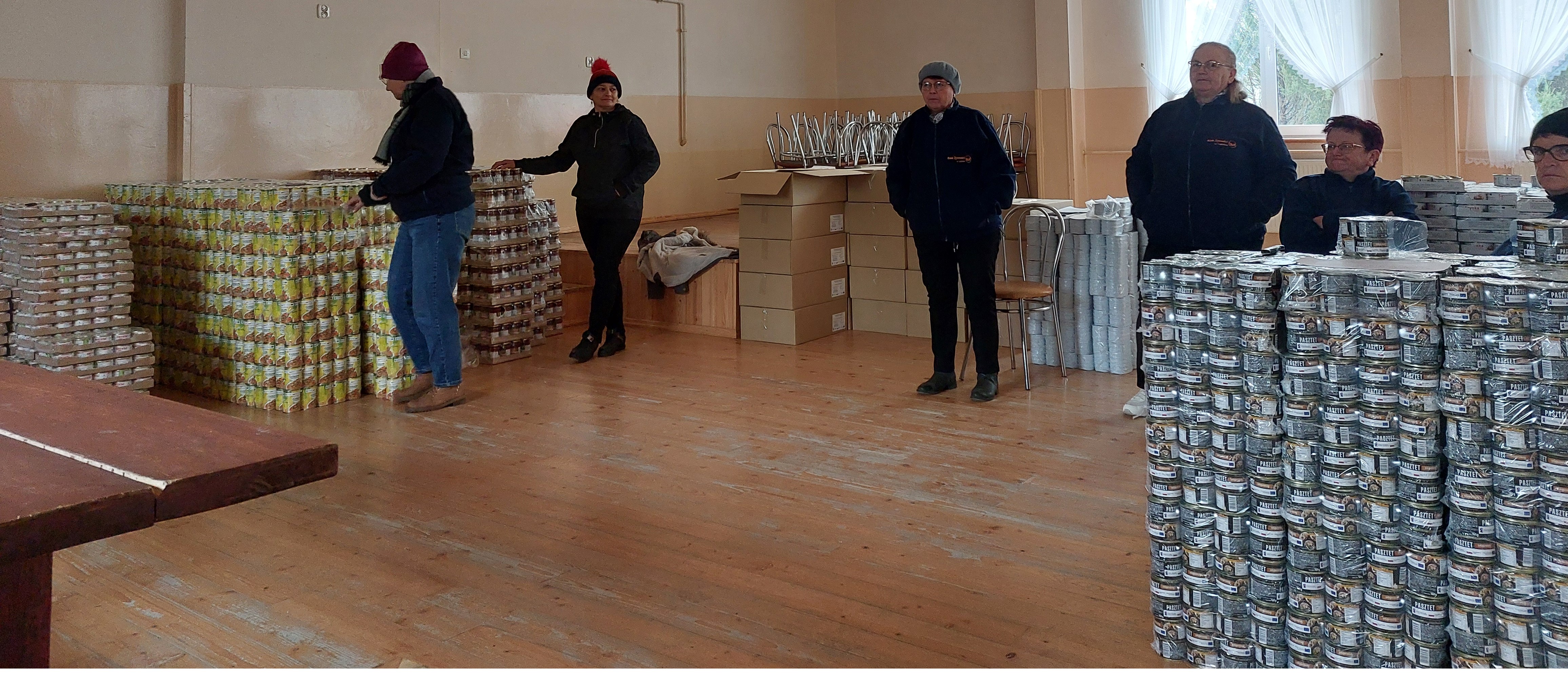 Pięć osób w ciemnych ubraniach i czapkach stoi w sali z drewnianą podłogą, wokół nich stosy puszek i kartonów, dwa okna z białymi zasłonami.
