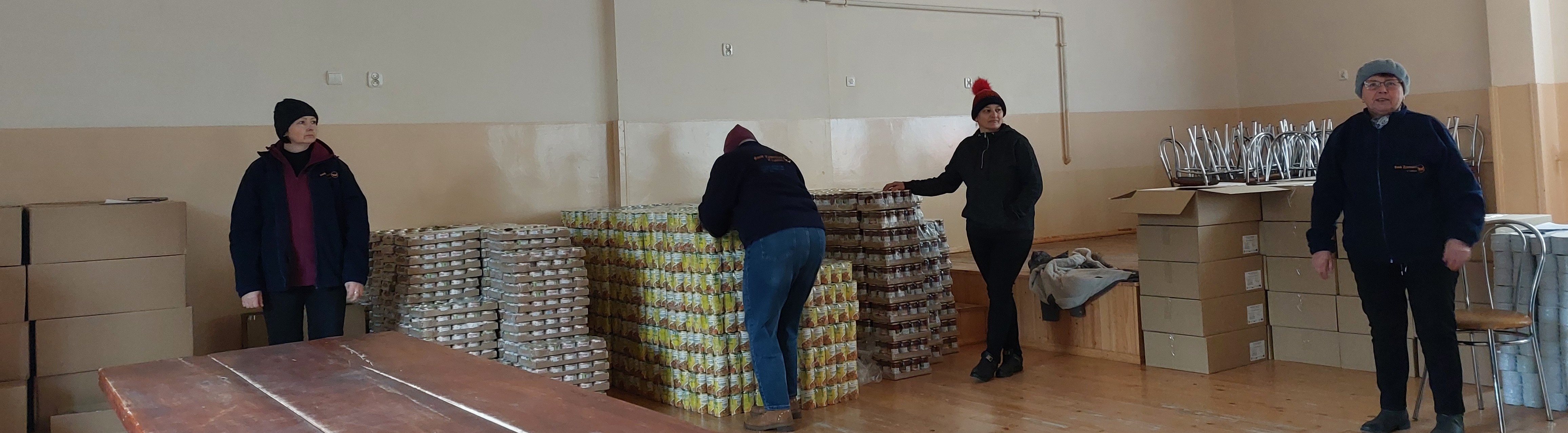 Cztery osoby w ciepłych ubraniach stoją i pracują przy stosach puszek i kartonów w jasnym pomieszczeniu z drewnianą podłogą.