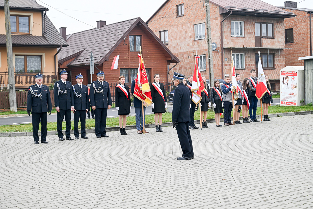 Grupa osób w mundurach i strojach formalnych stoi na placu z szarych kostek brukowych, trzymając czerwono-białe flagi, w tle drewniany i murowany dom.