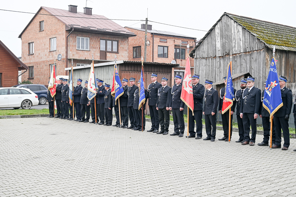 Grupa mężczyzn w mundurach strażackich stoi w rzędzie na szarym brukowanym placu, trzymając kolorowe flagi, w tle drewniane i murowane budynki oraz zaparkowane samochody.