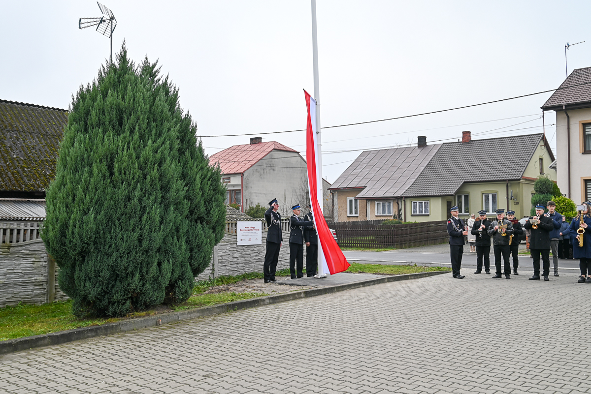 Grupa ludzi w mundurach przy masztach z biało-czerwoną flagą, domy z dachami i duże zielone iglaki przy chodniku z szarych kostek.