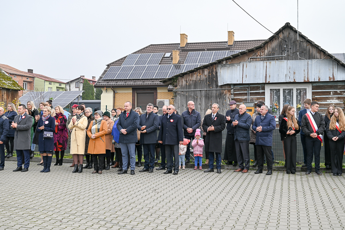 Grupa ludzi ubranych w płaszcze stoi na bruku przed domami z panelami słonecznymi na dachach, w tym dzieci i osoby z biało-czerwonymi kotylionami.