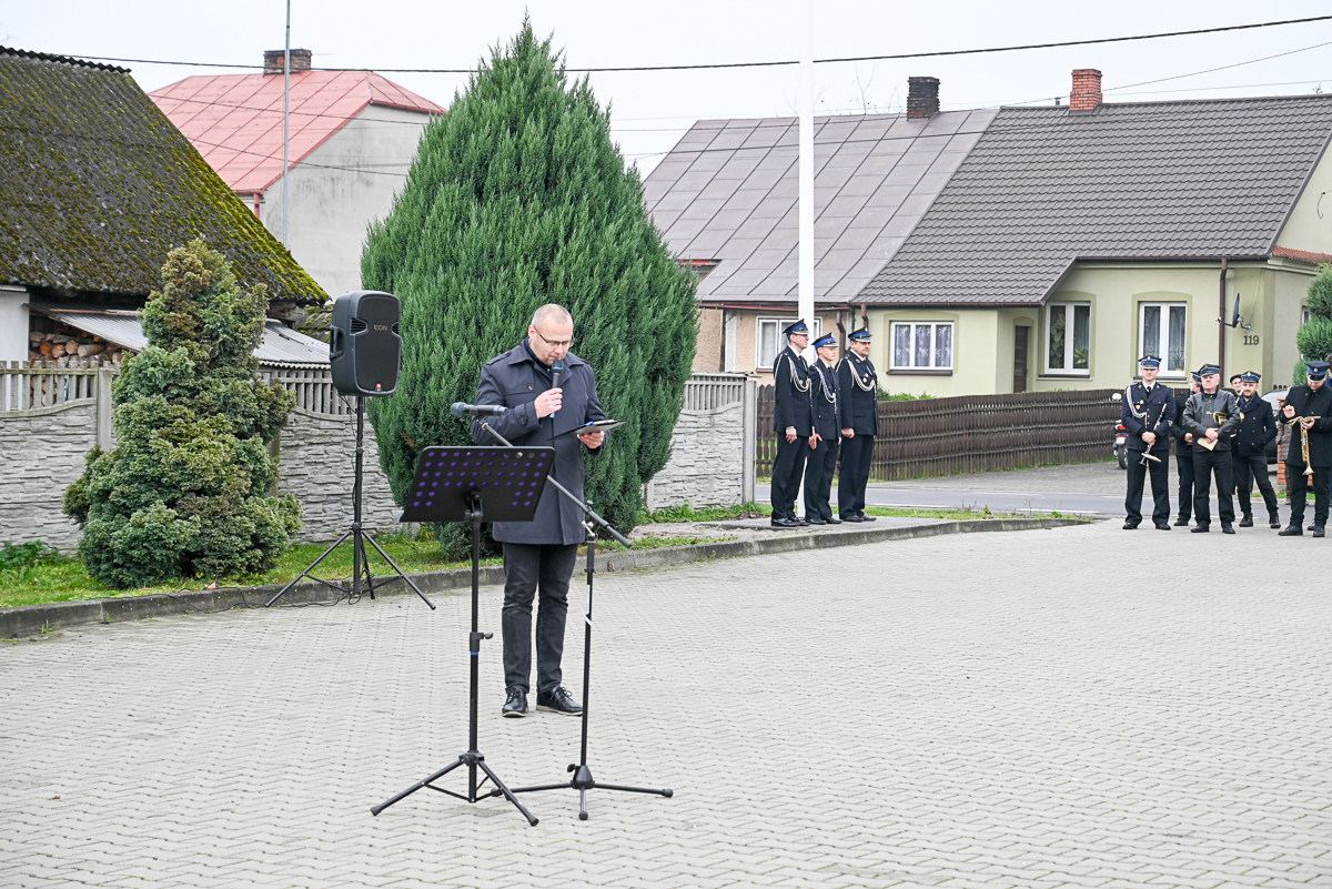 Mężczyzna w ciemnym płaszczu przemawia do mikrofonu na tle domów i zielonych drzew, obok stoi kilku umundurowanych mężczyzn, szara kostka brukowa, szary płot.