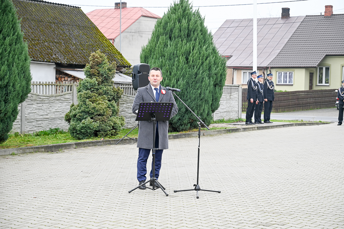 Mężczyzna w szarym płaszczu przy mikrofonie na stojaku na tle domów, zielonych drzew i trzech umundurowanych mężczyzn stojących z boku na chodniku z szarych kostek.