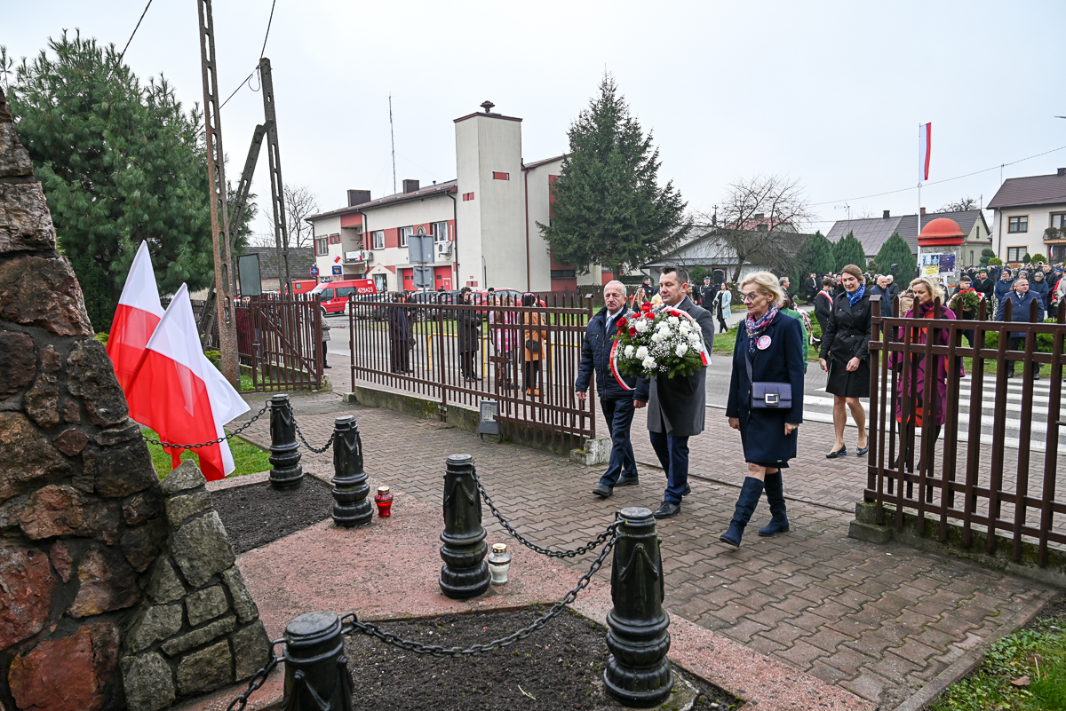 Kilka osób niesie biało-czerwony wieniec kwiatów na tle ogrodzenia, budynków i flag Polski przy pomniku z łańcuchami i zniczami.