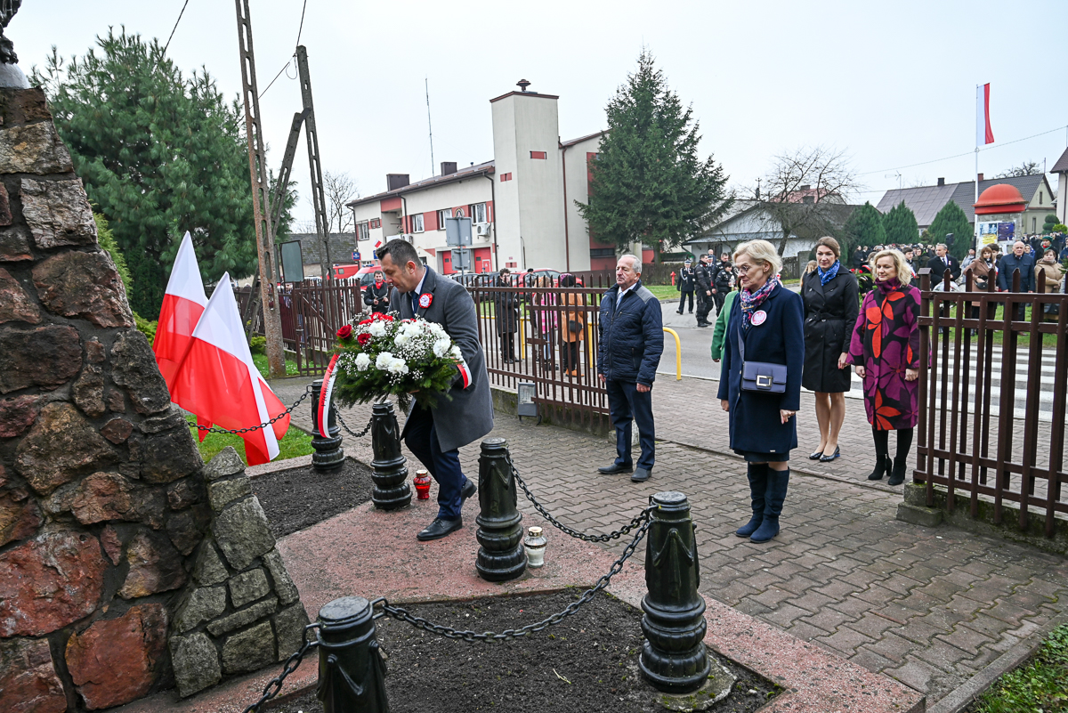 Mężczyzna w płaszczu z wieńcem kwiatów biało-czerwonych, kobiety i mężczyzna stojący na chodniku, biało-czerwone flagi, kamienna konstrukcja, metalowe łańcuchy.