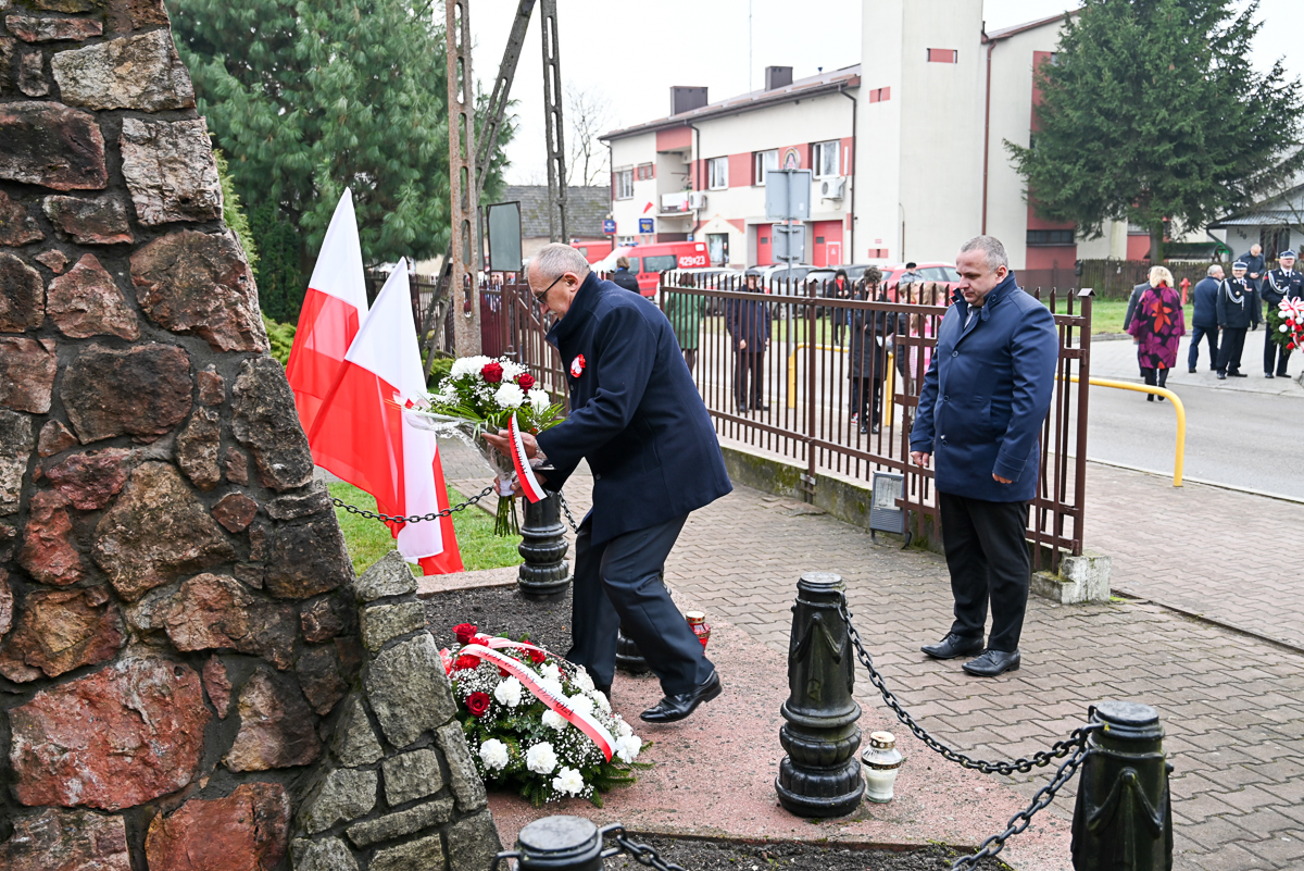 Mężczyzna w ciemnym płaszczu kładzie biało-czerwony wieniec kwiatów przy kamiennym pomniku, obok polskie flagi, drugi mężczyzna stoi obok, w tle płot i budynki.