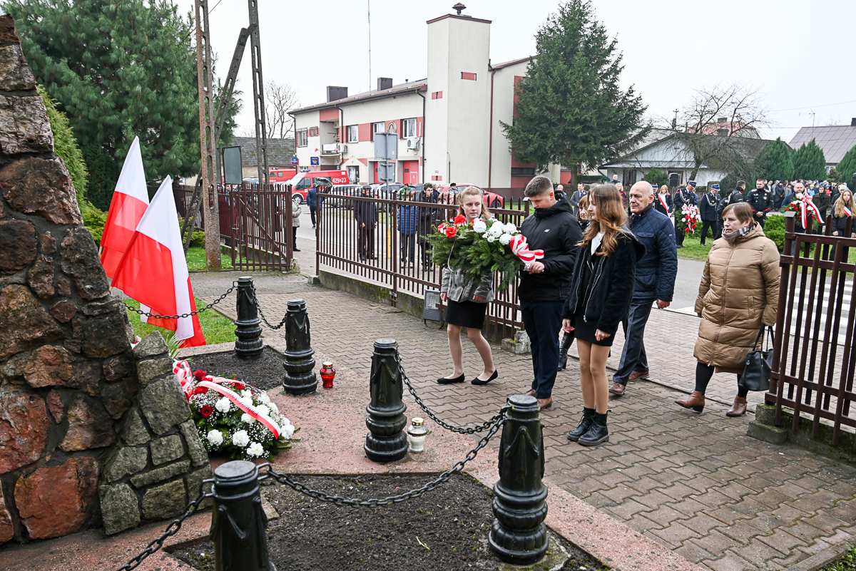Grupa ludzi w czarnych i beżowych ubraniach składa wiązankę biało-czerwonych kwiatów przy pomniku z kamienia, obok dwa biało-czerwone flagi, ogrodzenie i budynki w tle.