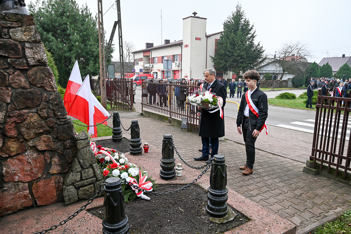 Mężczyzna i chłopiec z biało-czerwonymi szarfami składają wiązankę kwiatów biało-czerwonych przy kamiennym pomniku z łańcuchem i słupkami, w tle ludzie i flagi Polski.