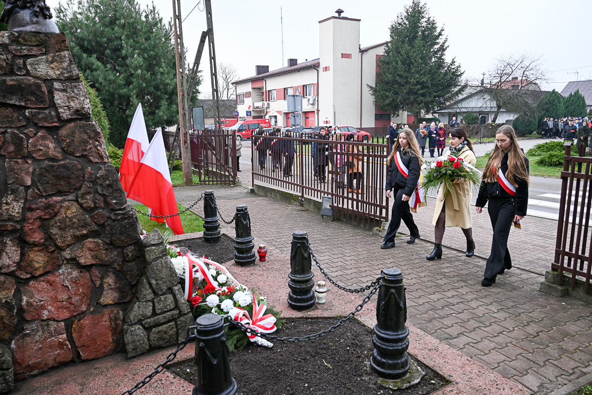 Pomnik z kamienia, biało-czerwone flagi, wieniec z kwiatów z biało-czerwoną wstążką, trzy kobiety z biało-czerwonymi szarfami niosące kwiaty, ludzie w tle, budynki i ogrodzenie.