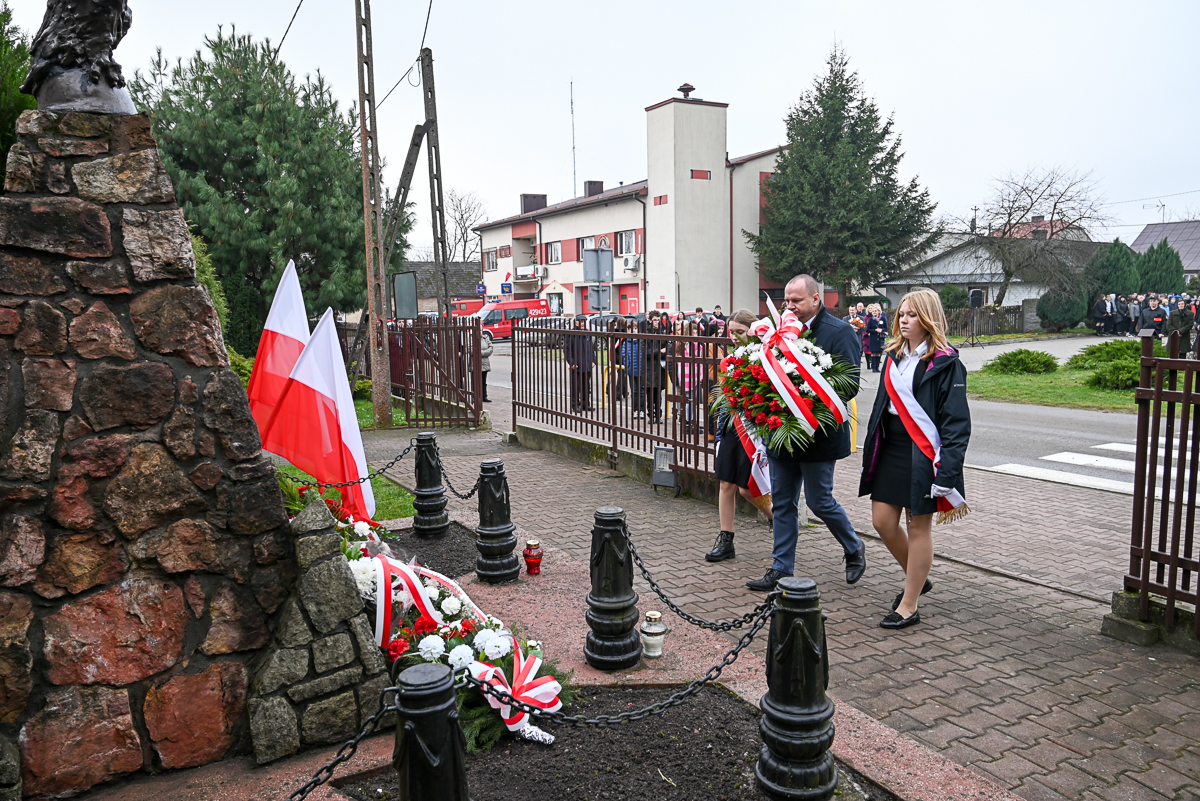 Pomnik z kamienia z biało-czerwonymi flagami, kwiaty z biało-czerwonymi wstążkami, trzy osoby niosące wieniec, w tle budynki i zgromadzeni ludzie.