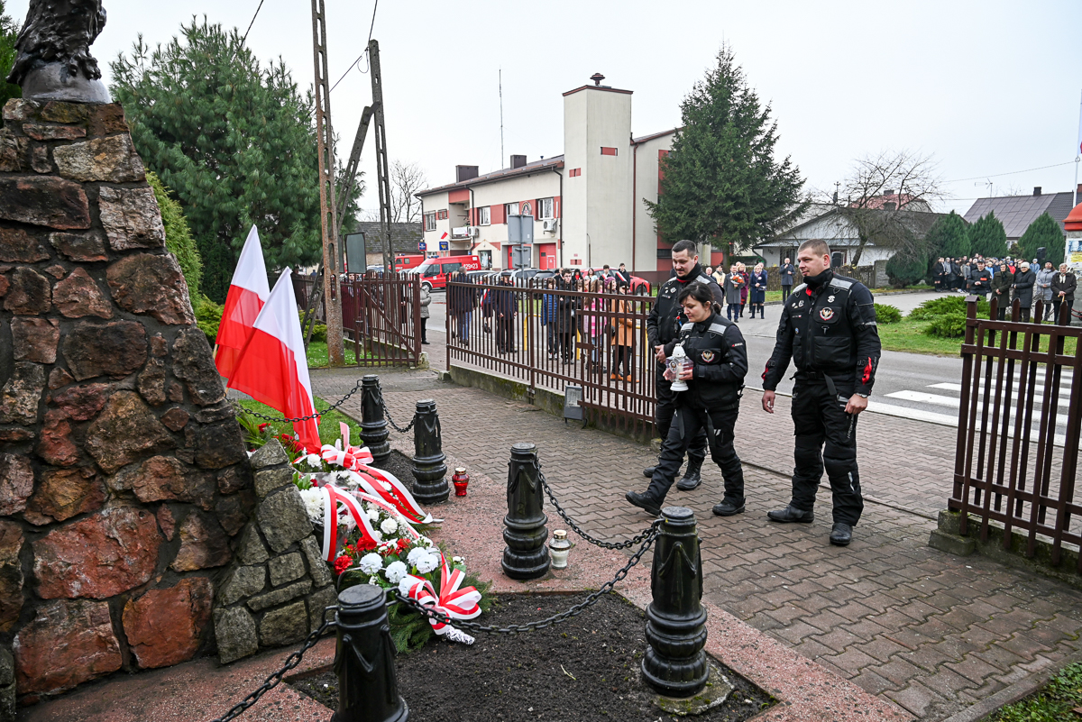 Pomnik z kamienia z biało-czerwonymi flagami i wieńcami, trzy osoby w czarnych mundurach niosące znicze, grupa ludzi za ogrodzeniem na tle budynków i drzew.