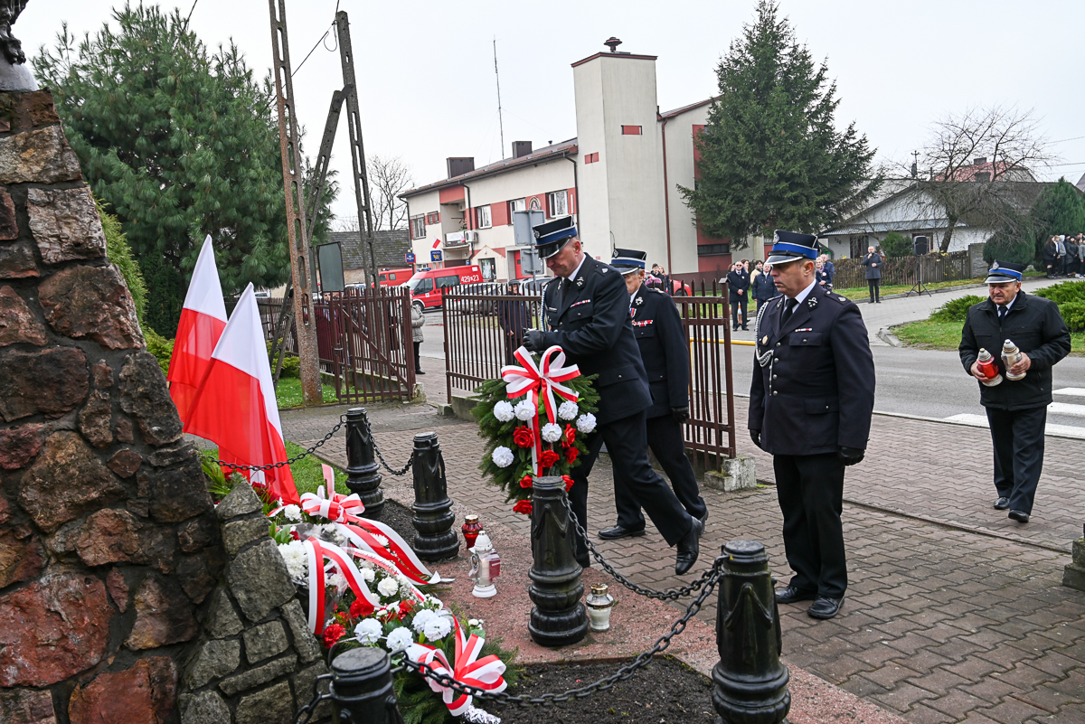 Czterech mężczyzn w mundurach składa wiązanki kwiatów przy pomniku z czerwono-białymi flagami i zniczami, w tle budynki i drzewa na pochmurnym niebie.