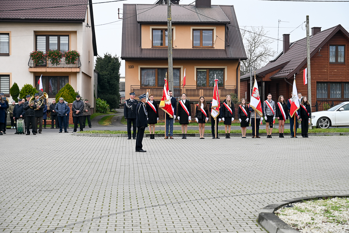 Grupa ludzi w mundurach i strojach oficjalnych z biało-czerwonymi flagami, stojąca na szarej brukowanej nawierzchni, w tle domy mieszkalne i widoczne elementy zieleni.