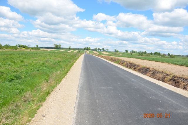 Asfaltowa droga między zielonymi polami pod niebieskim niebem z białymi chmurami.