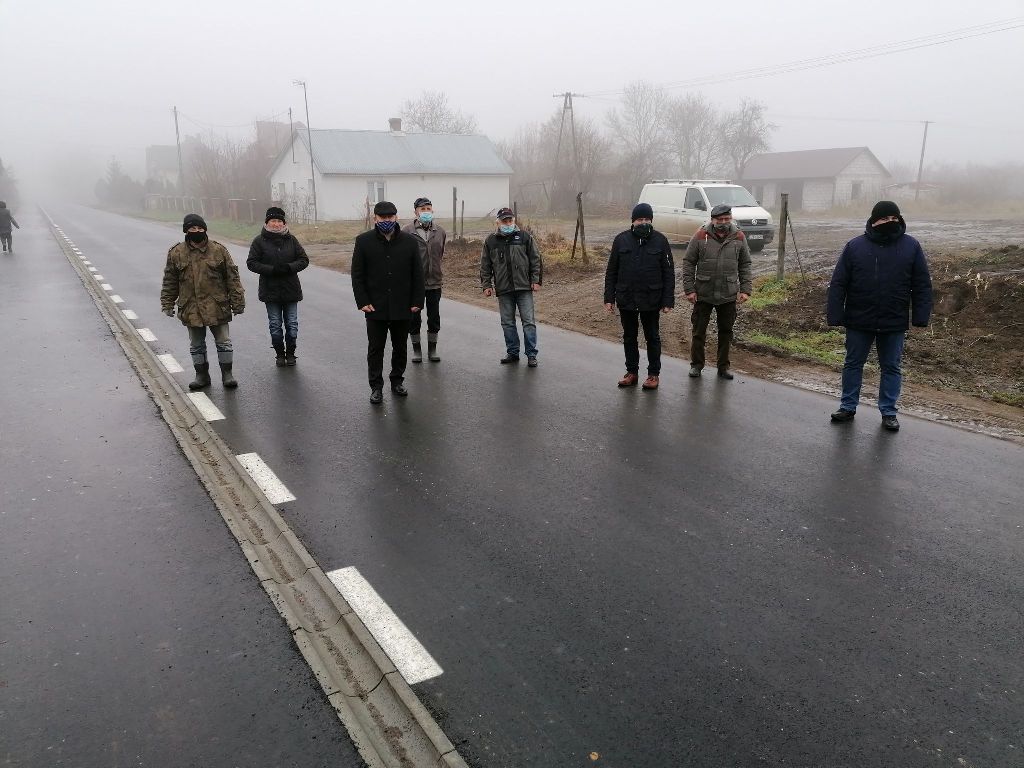 Osiem osób w kurtkach i maseczkach stoi na mokrej, pustej drodze przy mglistym, szarym niebie, w tle białe domy i biały van.