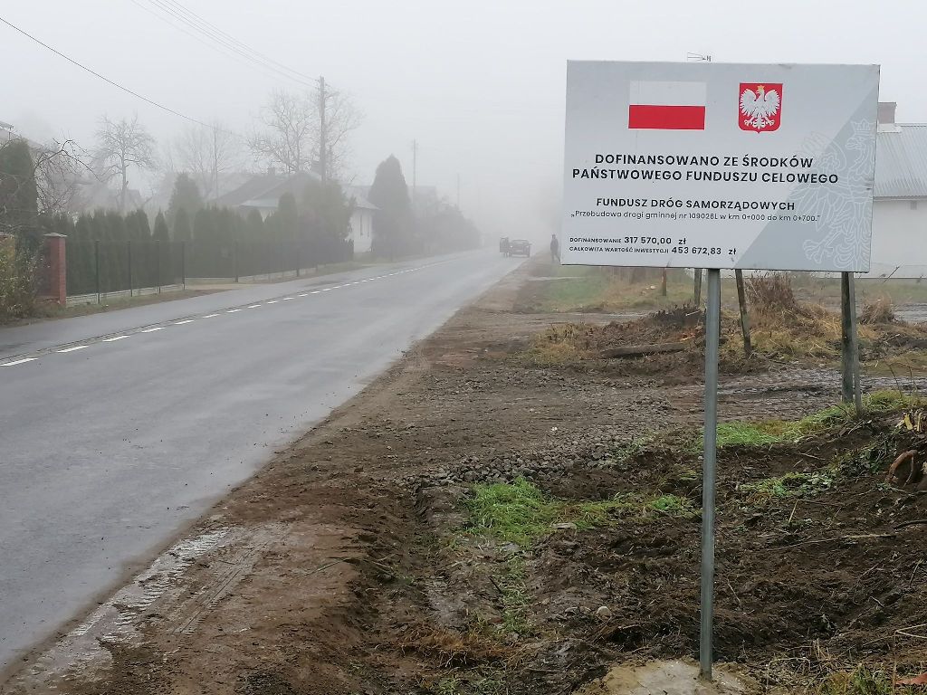 Droga asfaltowa w mglisty dzień, po prawej tablica informacyjna z flagą Polski i godłem, obok ziemia i trawa, w tle domy i drzewa.