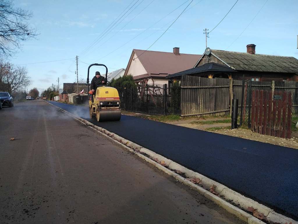 Żółty walec drogowy na świeżo położonym czarnym asfalcie obok drewnianych ogrodzeń i domów pod niebieskim niebem.
