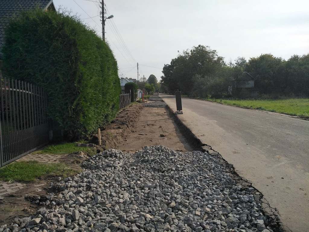 Budowa chodnika z kamieni i ziemi wzdłuż asfaltowej drogi, po lewej zielone żywopłoty i ogrodzenia, po prawej trawnik i drzewa.