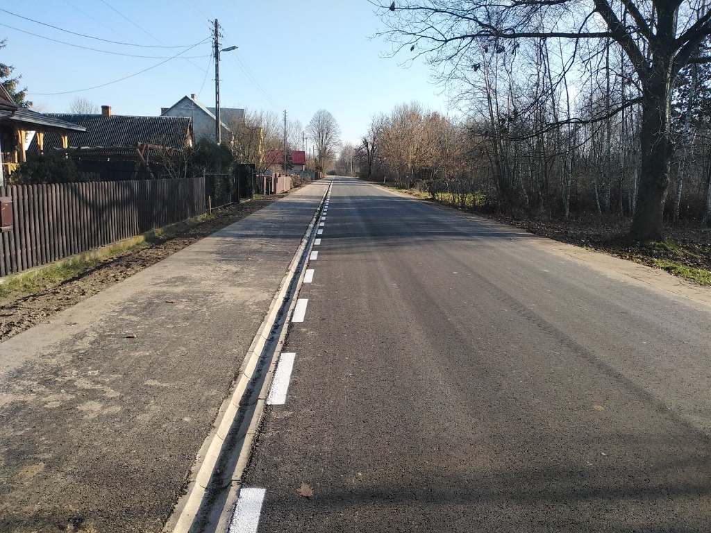 Asfaltowa droga z białymi liniami, po lewej stronie chodnik, po lewej drewniane ogrodzenia i domy, po prawej drzewa bez liści.