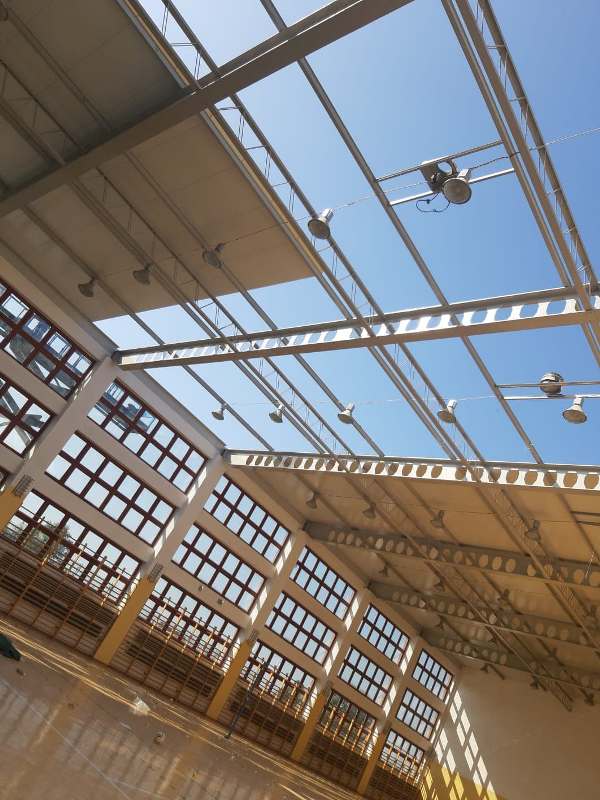 Wnętrze hali sportowej z dużymi oknami, metalową konstrukcją sufitu, licznymi lampami i drewnianą podłogą odbijającą światło.