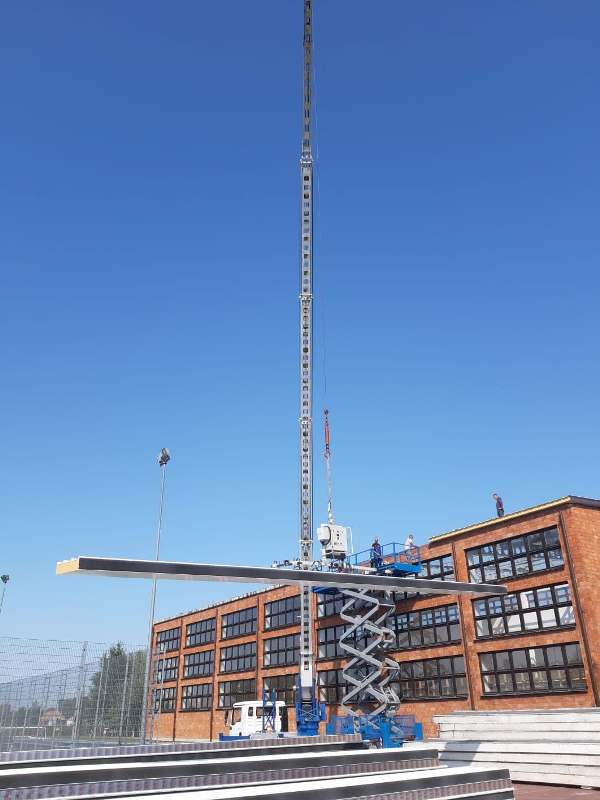 Duży dźwig podnoszący długi stalowy element nad placem budowy obok ceglanego budynku z wieloma oknami, pod nim platforma nożycowa z dwoma osobami.