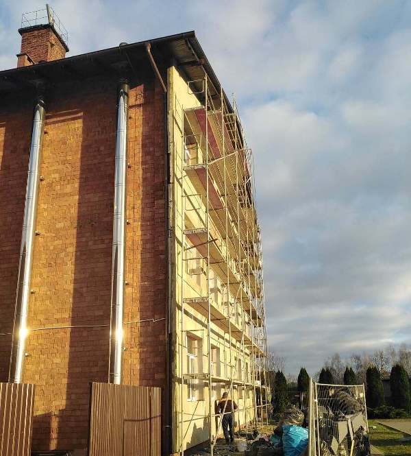 Budynek z czerwoną cegłą i żółtą elewacją częściowo osłonięty rusztowaniem, niebo z chmurami, na pierwszym planie ogrodzenie i materiały budowlane.