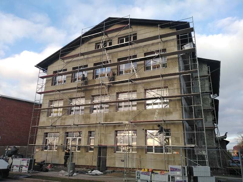 Trójkondygnacyjny budynek z rusztowaniem, na którym pracuje kilka osób, szara elewacja, okna z brązowymi ramami, niebo z chmurami.