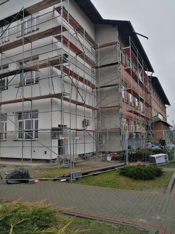 Budynek z rusztowaniem, biała i czerwona elewacja, ogrodzenie budowlane, chodnik i trawniki, pochmurne niebo.