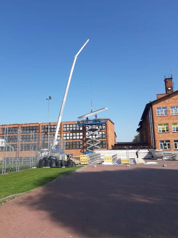 Czerwone ceglane budynki, dźwig podnoszący belkę, podnośnik nożycowy z ludźmi na dachu, stosy białych i żółtych materiałów budowlanych, ogrodzenie i zielona trawa.