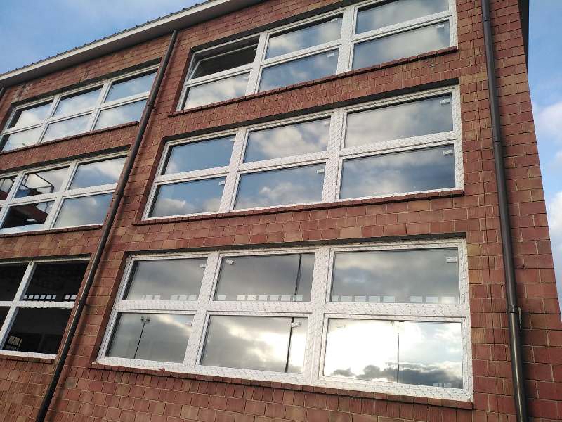 Budynek z czerwonej cegły z trzema rzędami dużych okien z białymi ramami, odbijające niebieskie niebo z chmurami.
