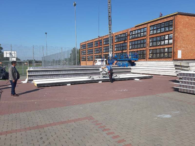 Czerwony budynek z dużymi oknami, na pierwszym planie betonowe płyty, mężczyźni, niebieska maszyna do podnoszenia, ogrodzenie boiska i niebieskie niebo.