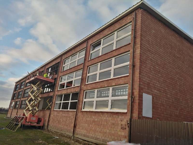 Ceglany budynek z trzema rzędami białych okien, podnośnik koszowy z dwoma osobami, częściowo zachmurzony niebieski nieboskłon.