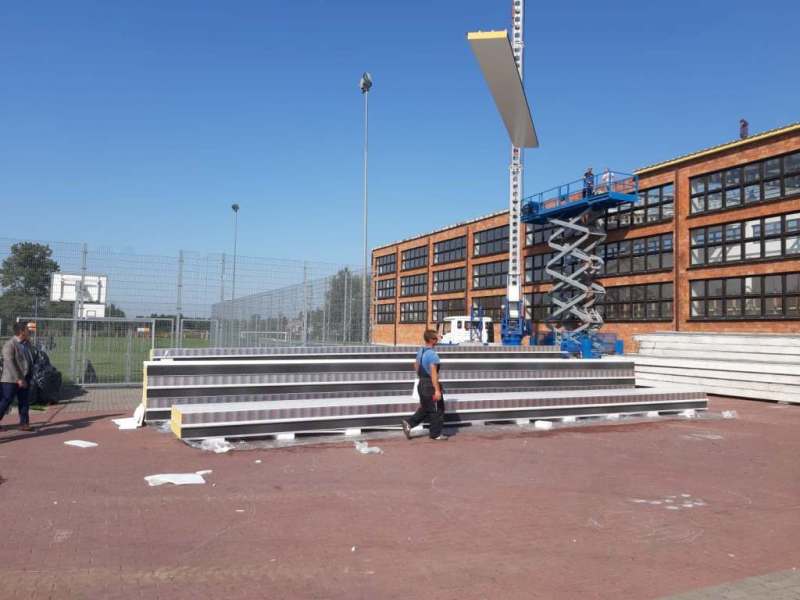 Stosy białych paneli na zewnątrz, dźwig podnosi panel, dwóch mężczyzn, budynek z czerwonej cegły i ogrodzenie z metalowej siatki.